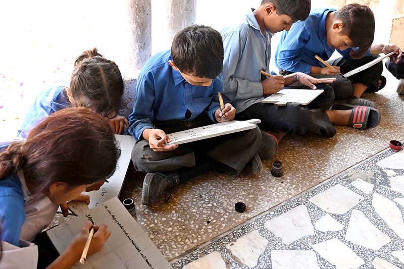 Students are learning writing skills through the traditional old method using ink and wooden pens (qalam) on wooden writing boards.