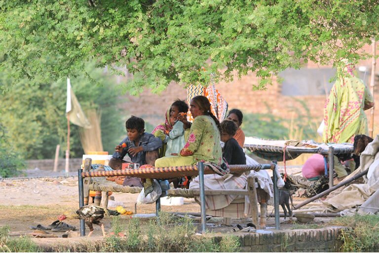 A nomadic family resting on woven cots beneath a tree along a canal bank on the city’s outskirts