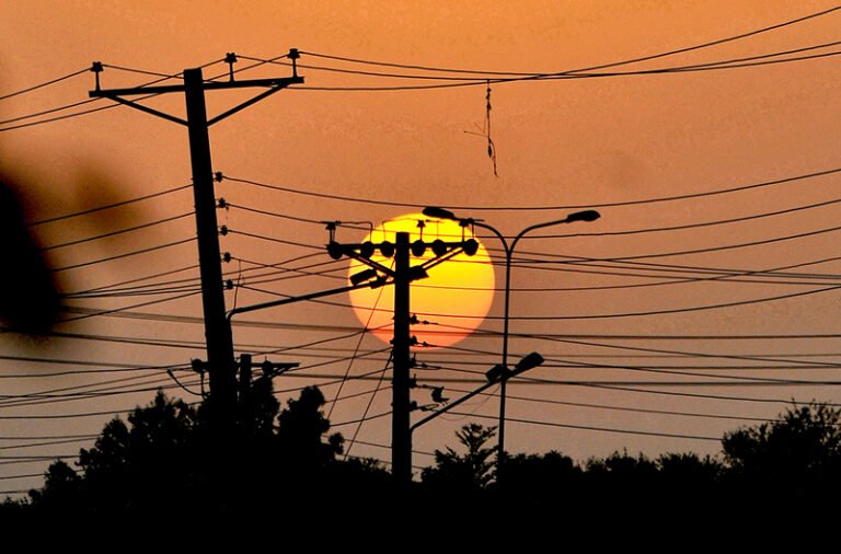 The setting sun creates a scenic silhouette of electricity transmission poles and wires against the evening sky.