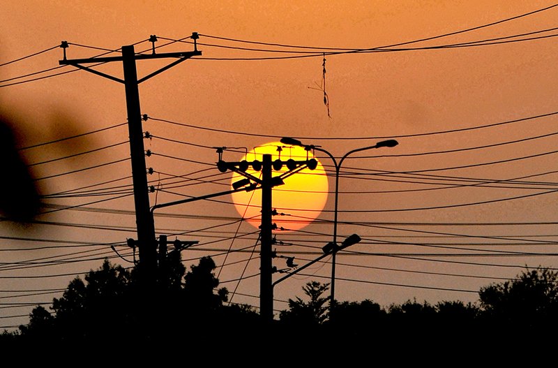 The setting sun creates a scenic silhouette of electricity transmission poles and wires against the evening sky.