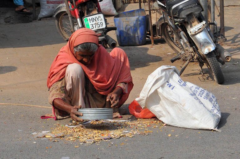 An elderly woman collects scattered grains from the roadside into a sack near a grain shop in the city