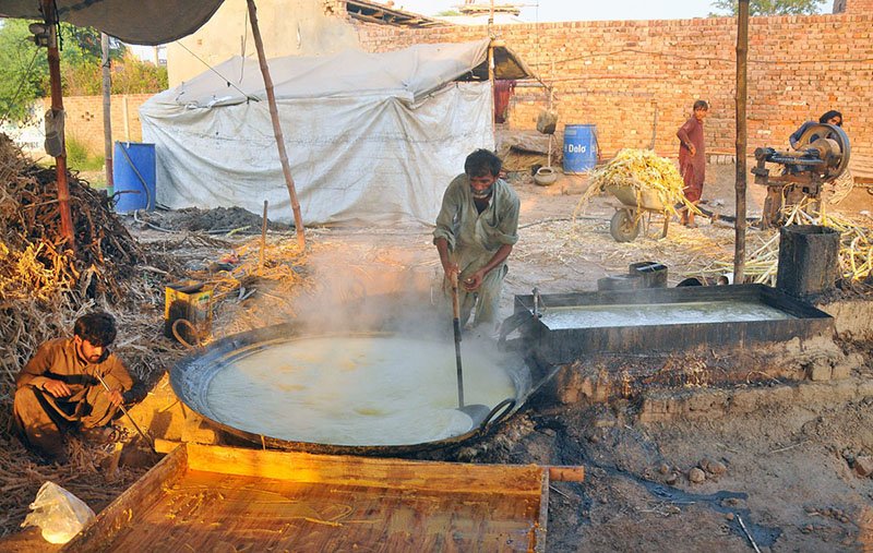 A worker boils sugarcane juice in a large pan while another tends the furnace fire to prepare traditional gur (jaggery) at a roadside setup in the city